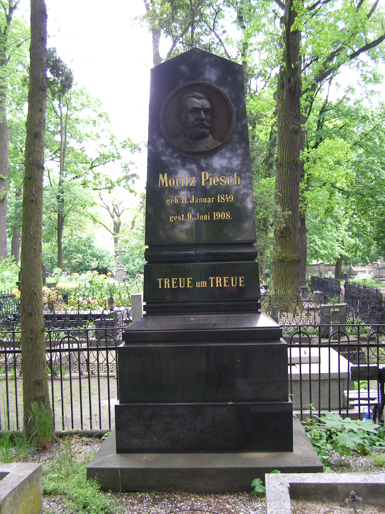Czarny granitowy obelisk z okrągłą płaskorzeźbą męskiej głowy, pod nią złocone napisy: imię i nazwisko, daty urodzenia i śmierci po niemiecku oraz „Treue um Treue”, wokół drzewa i kute ogrodzenia sąsiednich grobów